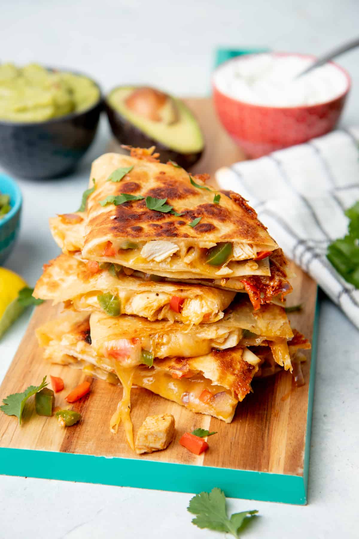 A stack of quesadillas sits on a wooden cutting board. A red bowl of sour cream sits in the background.