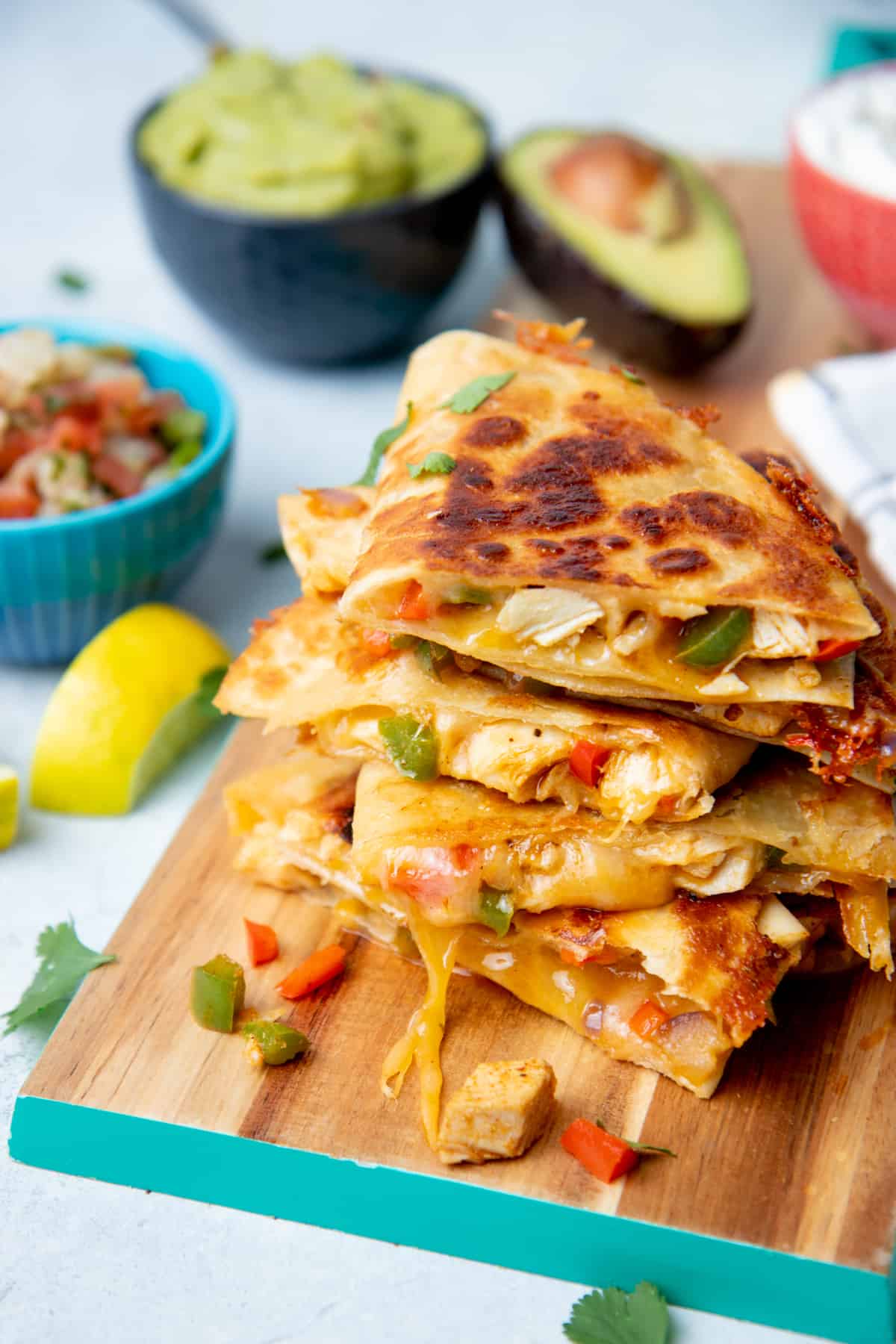 Chicken quesadillas rest on a wooden cutting board. A bowl of salsa and a bowl of guacamole sit behind them.