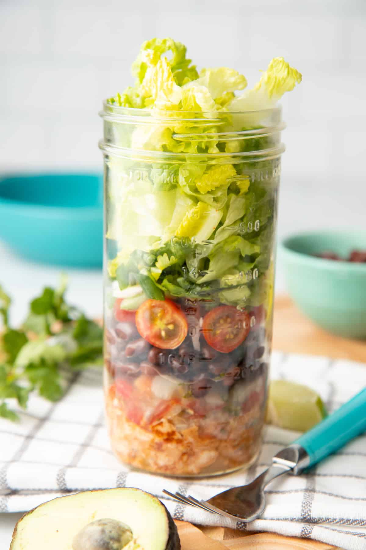 A mason jar layered with components for a chicken taco salad sits on a folded dish towel, surrounded by ingredients.