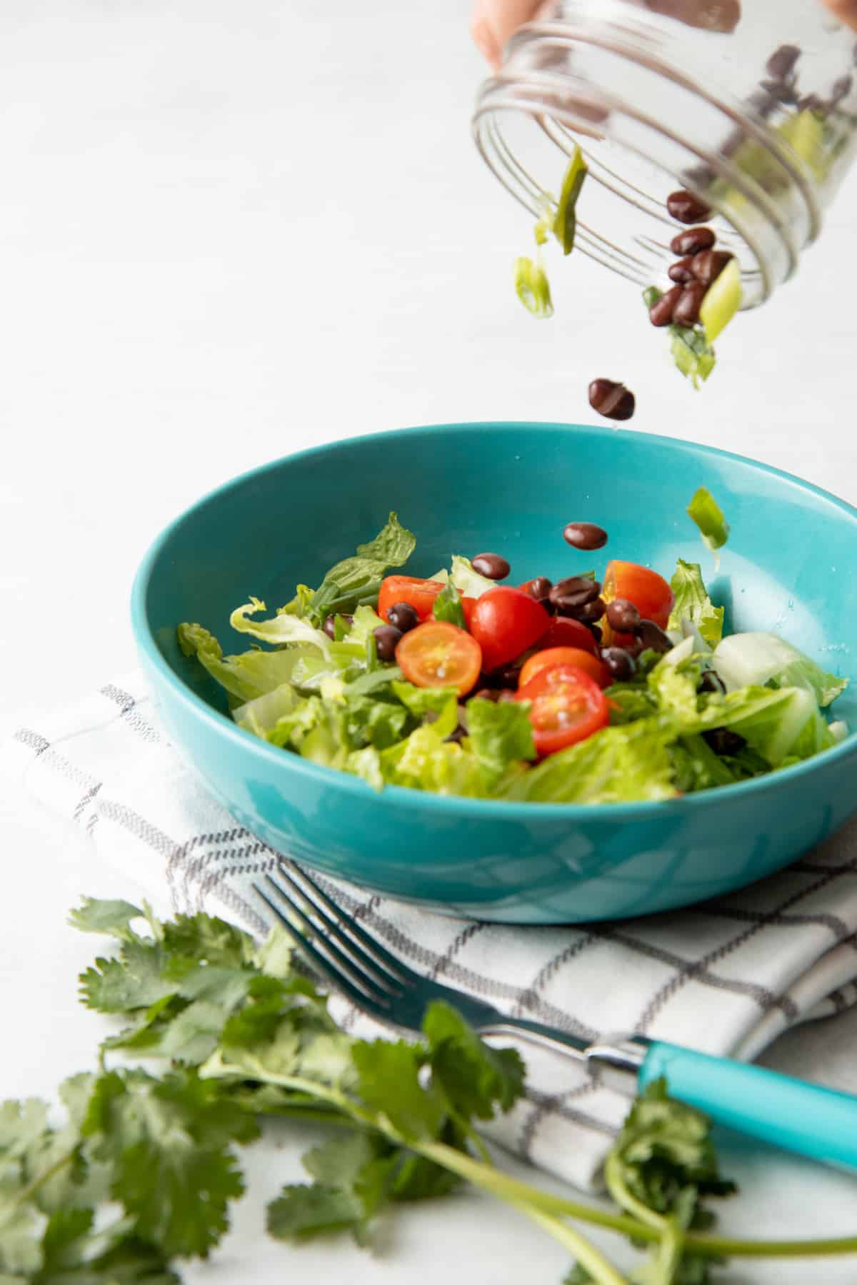 Black beans and lettuce pour out of a jar into a teal bowl. The bowl is filled with lettuce, tomatoes, and black beans.