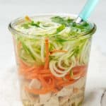 A small jar is filled with chicken zoodle soup. A spoon with a teal handle sits in the jar.