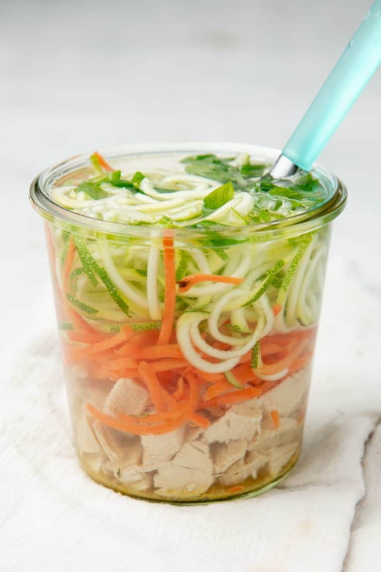 A small jar is filled with chicken zoodle soup. A spoon with a teal handle sits in the jar.