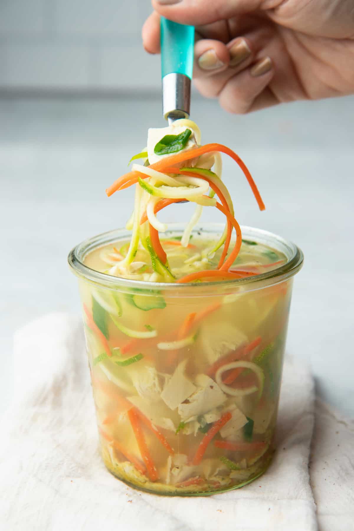 A hand holds a spoonful of chicken zoodle soup. A jar of soup sits underneath.