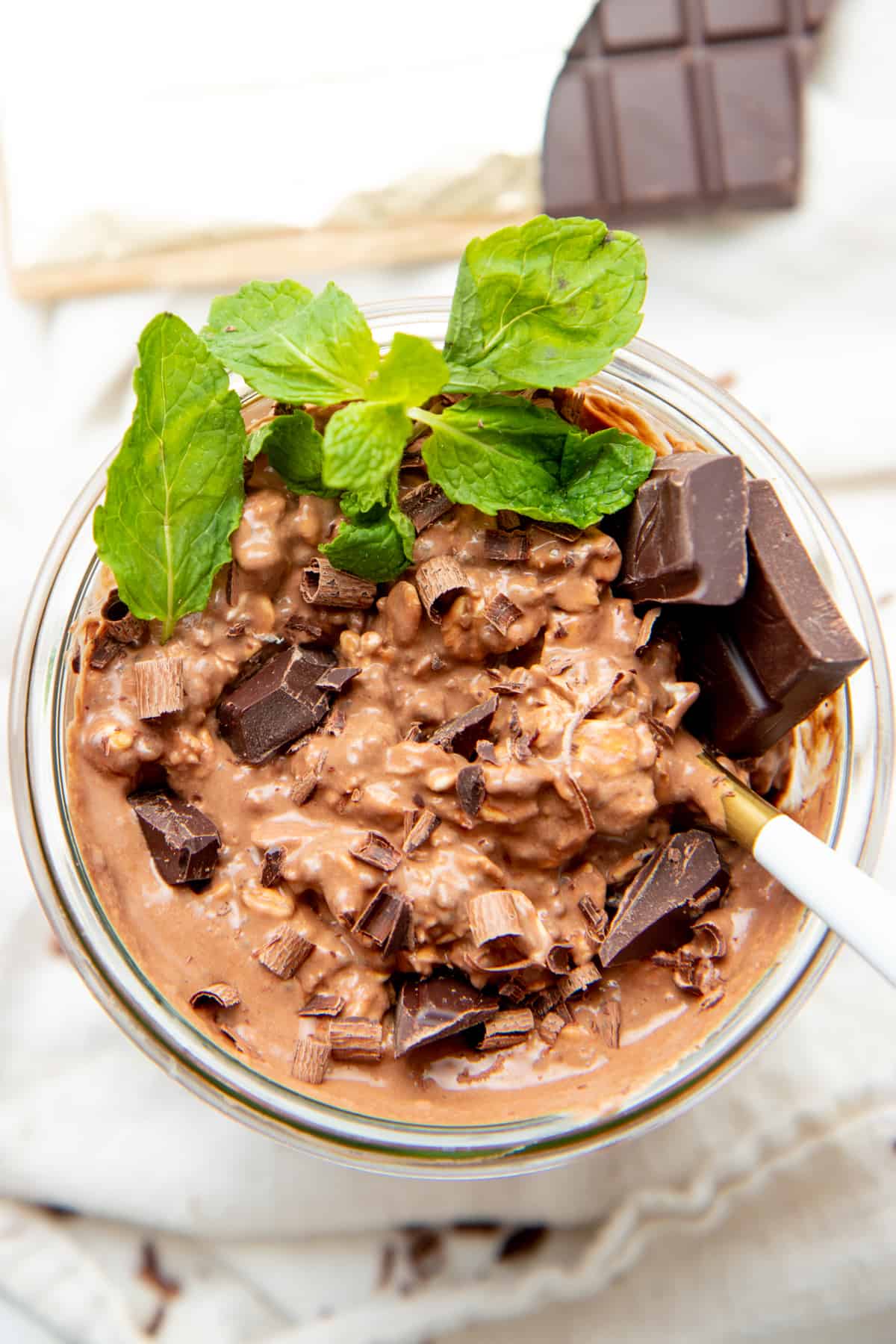 A spoon rests in a jar of overnight oats. Oats are topped with chopped chocolate and mint leaves.
