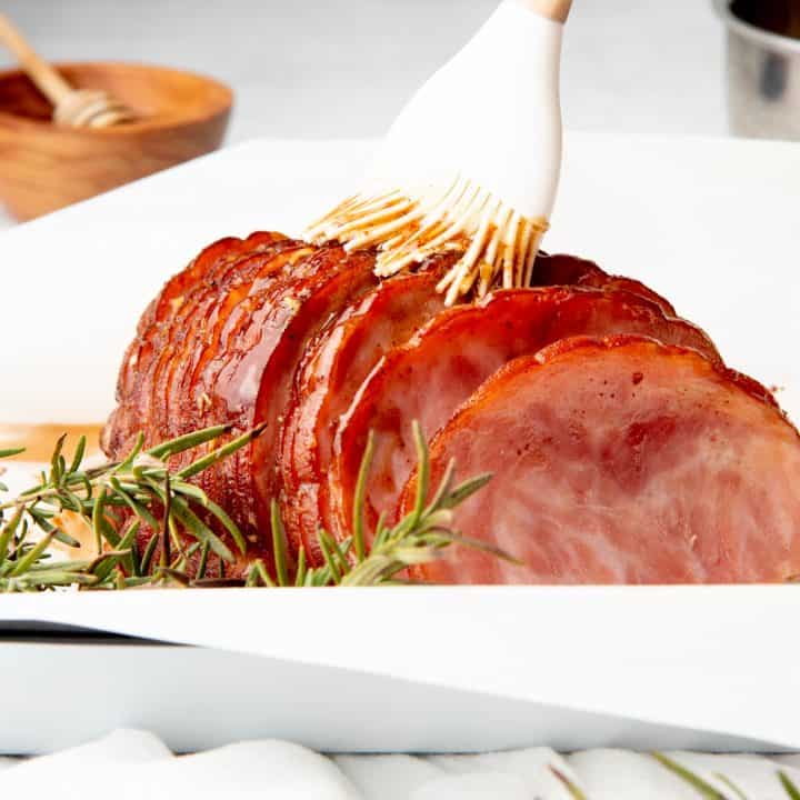 A hand bastes ham with a white brush. The dish sits on a white tablecloth.