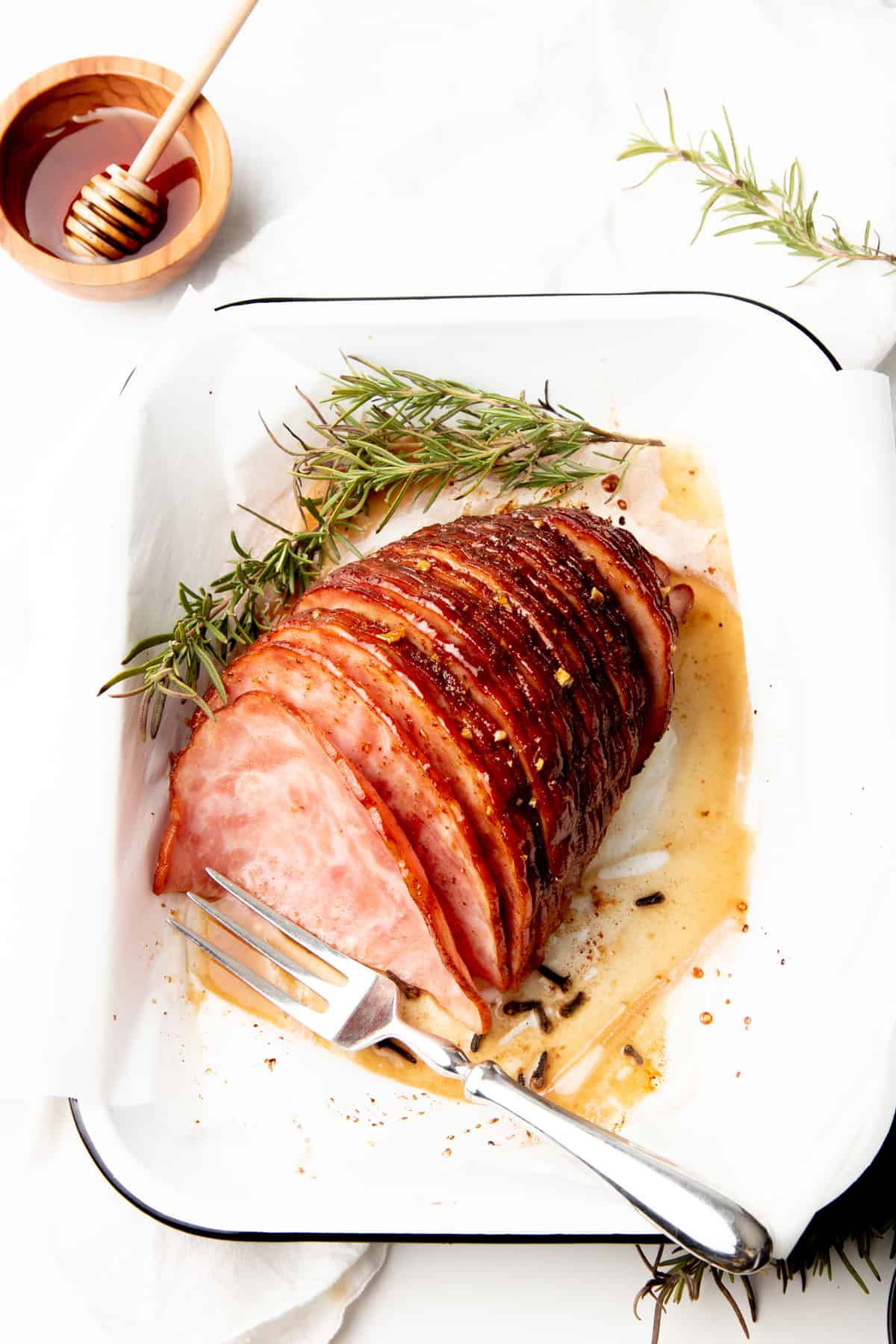 Sliced ham sits in a white rectangular dish. A small bowl of honey is on the side.