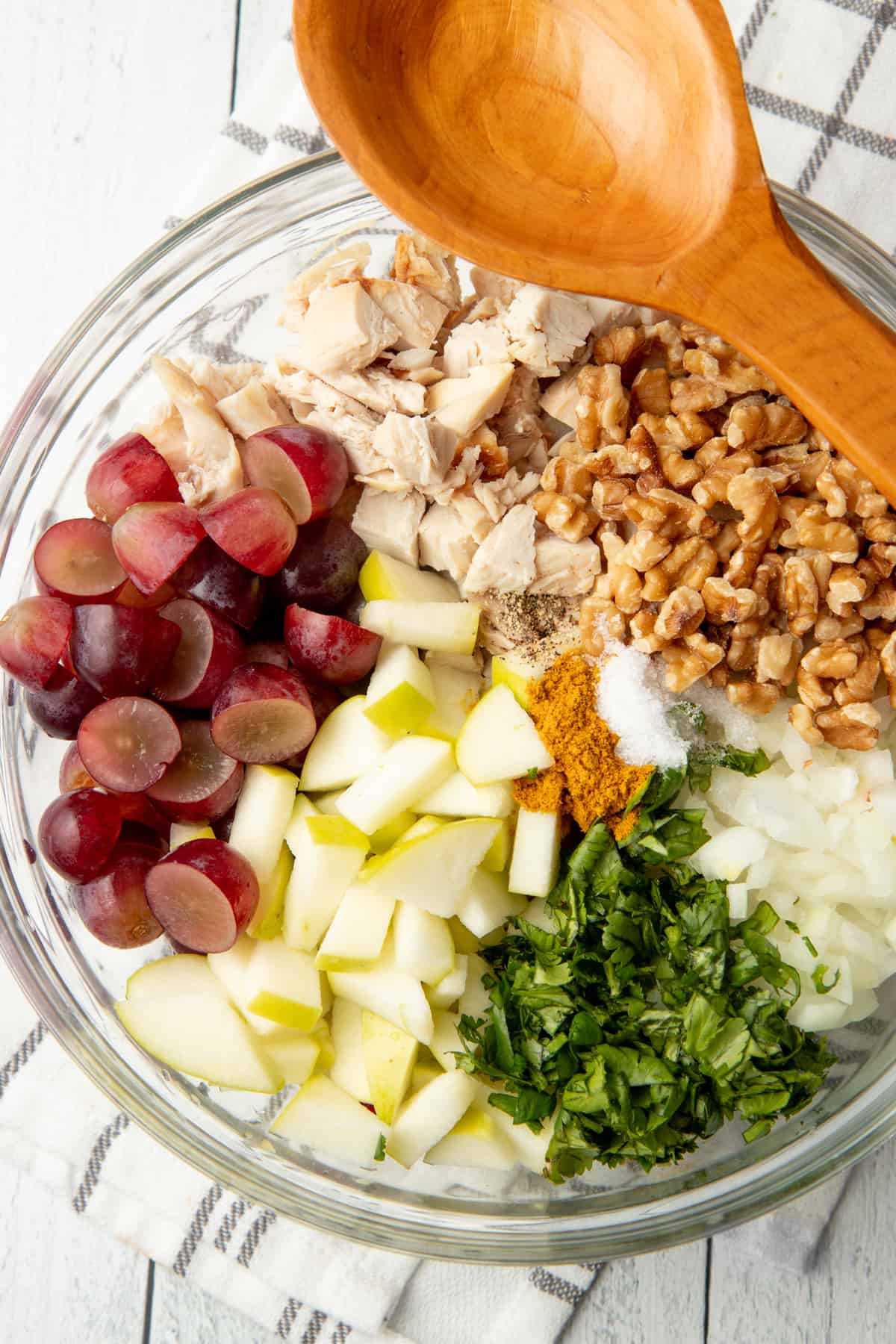 Grapes, chopped chicken, walnuts, onion, and apples are in a glass bowl. A wooden spoon sits on top of the bowl.