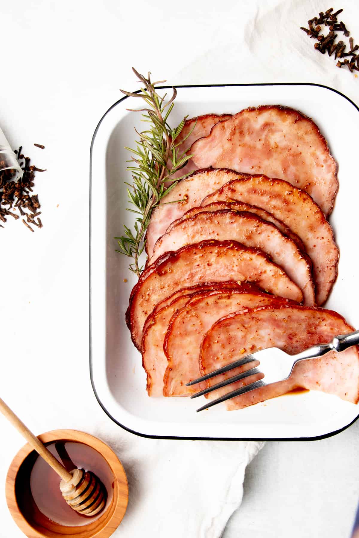 Sliced slow cooker ham rests in a white dish. A sprig of rosemary sits in the top left corner of the dish.