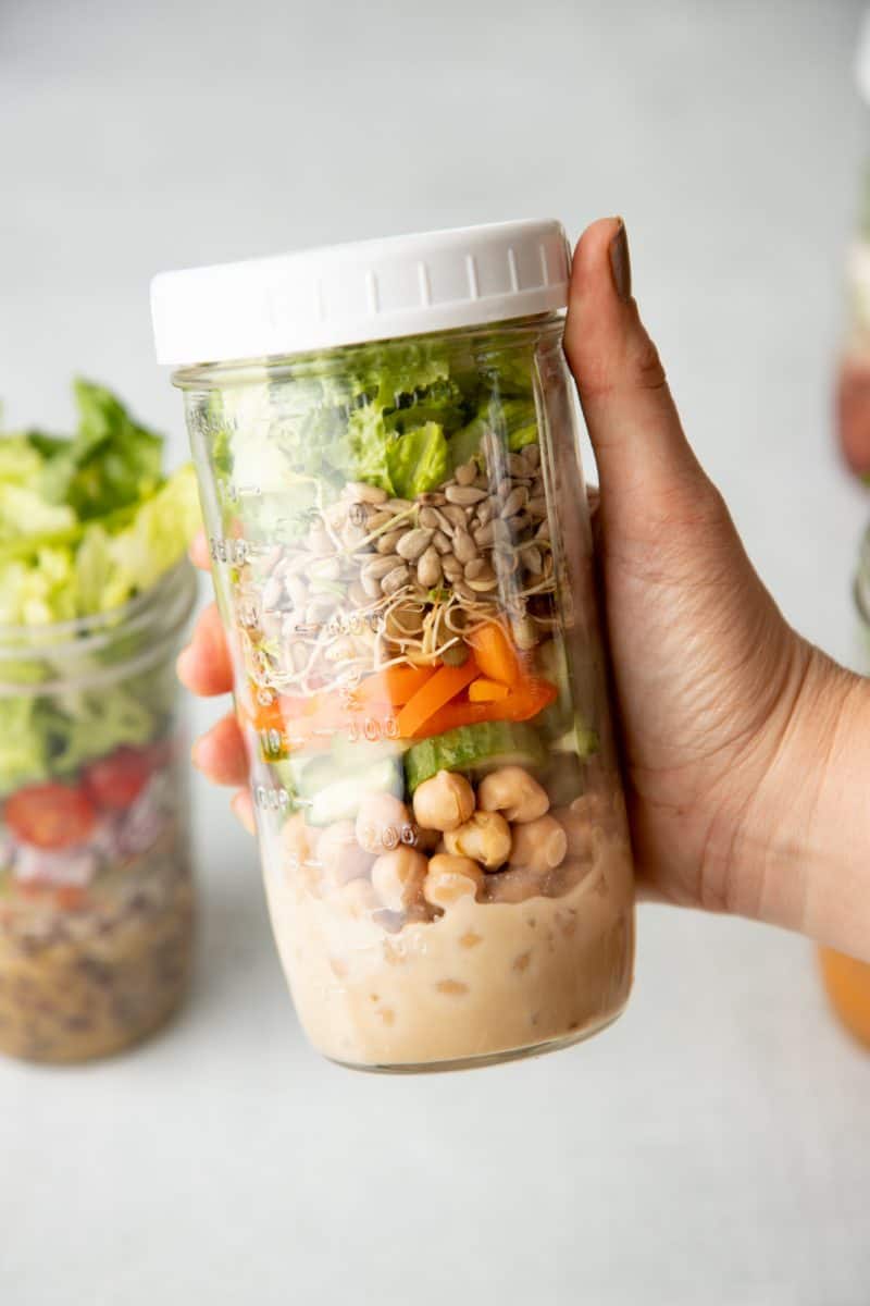 A hand holds a tall mason jar with a white lid. Jar is layered with salad ingredients, including dressing, chickpeas, vegetables, seeds, and lettuce.