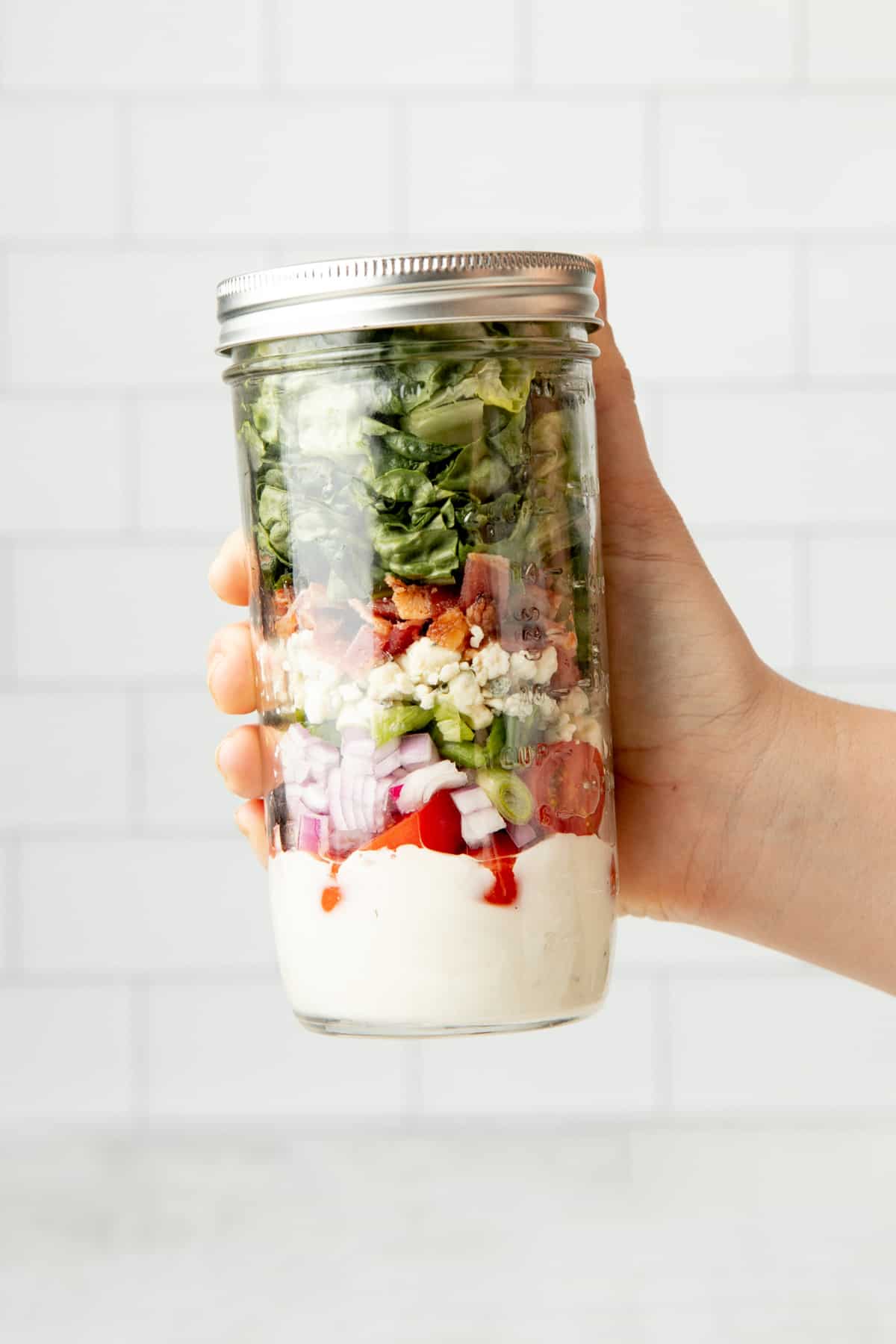A hand holds a capped mason jar filled with the components of a wedge salad.