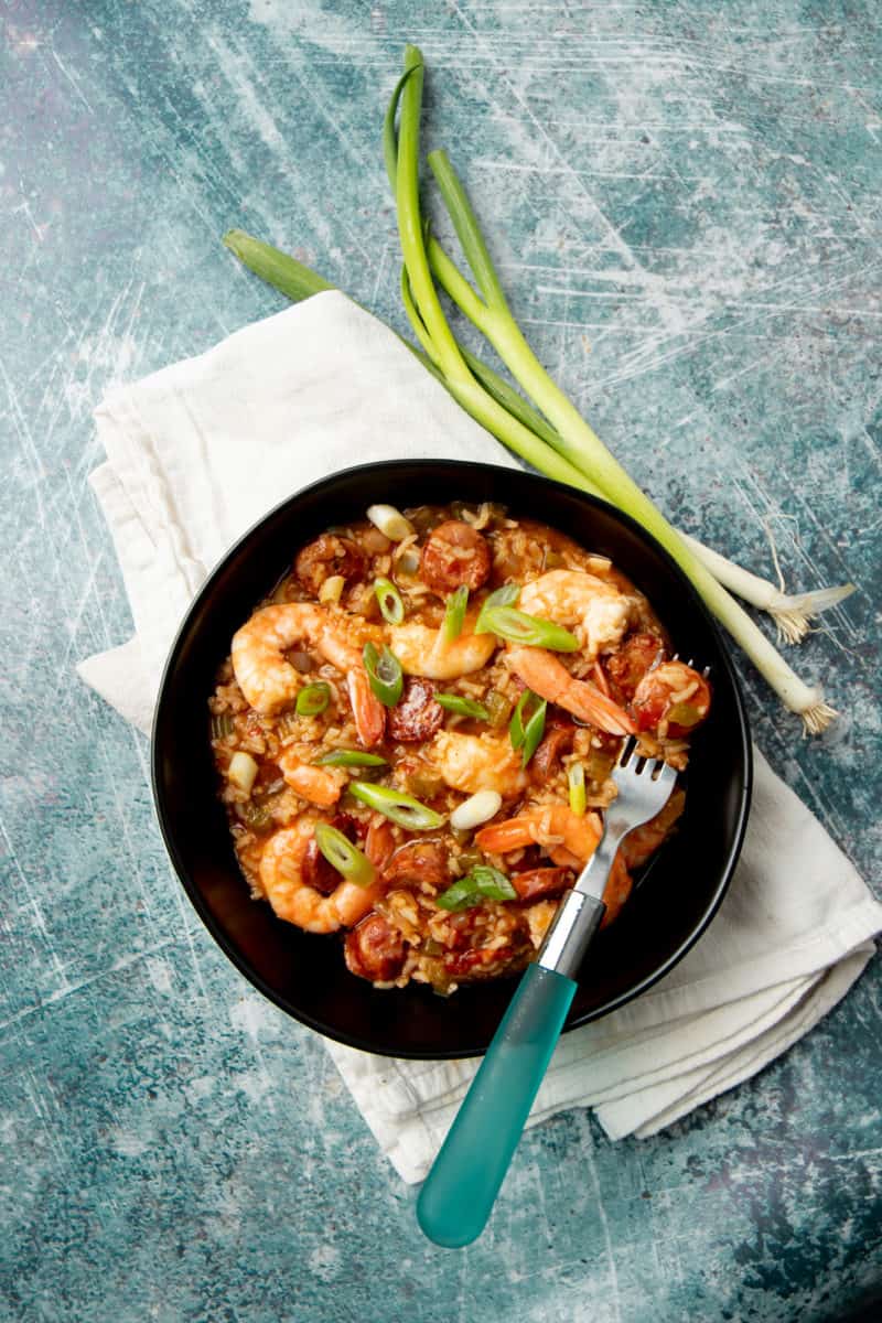 A piece of sausage rests on a fork inside a bowl of jambalaya. Whole green onions lay alongside the bowl.