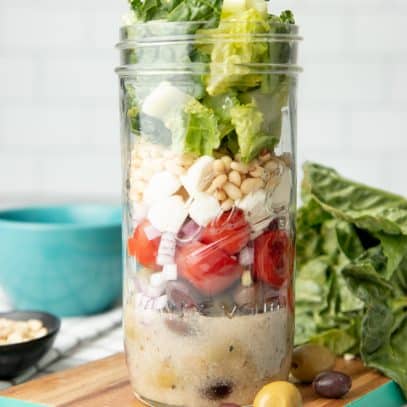 A mason jar salad sits on a teal-edged cutting board, surrounded by lettuce, olives, and cherry tomatoes.