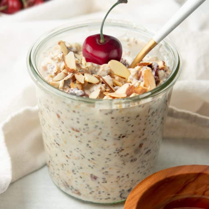 A spoon rests in a glass jar of cherry and almond overnight oats. The oats are garnished with a whole fresh cherry.