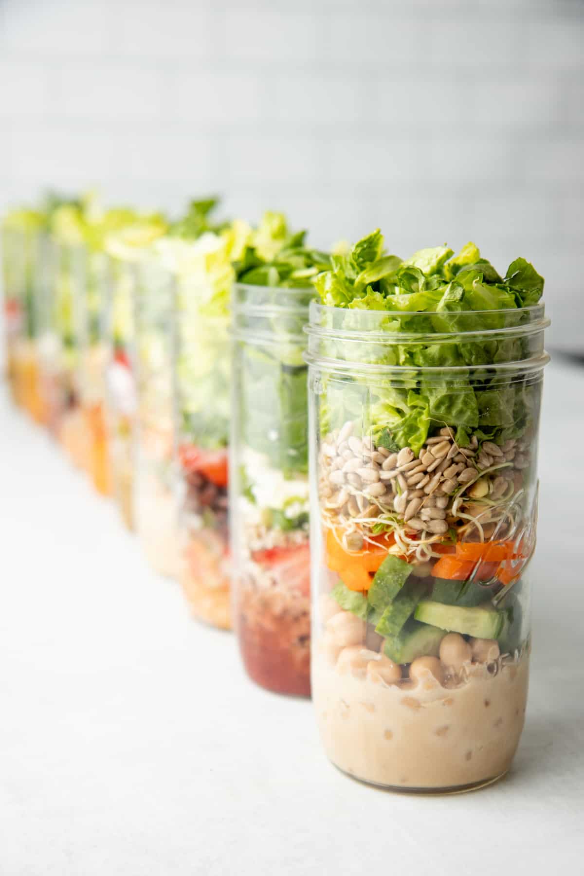 Ten tall mason jars are layered with salad ingredients.