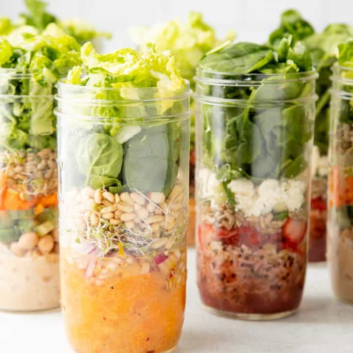Tall mason jars are layered with different salads. Jar in front includes mandarin oranges, sprouts, and lettuces.