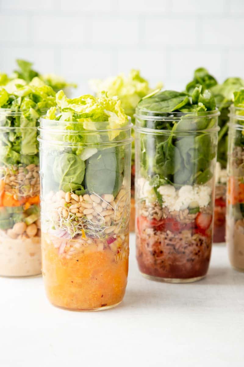 Tall mason jars are layered with different salads. Jar in front includes mandarin oranges, sprouts, and lettuces.