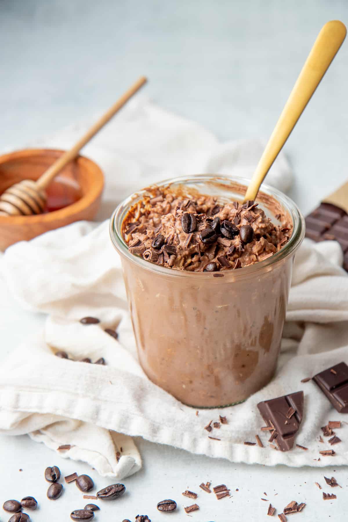 A gold spoon rests in a jar of chocolate overnight oats. A small bowl of honey also sits on the white dishtowel.