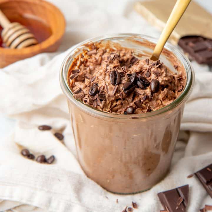 A gold spoon sits in a jar full of mocha overnight oats. The jar rests on a white dishtowel.