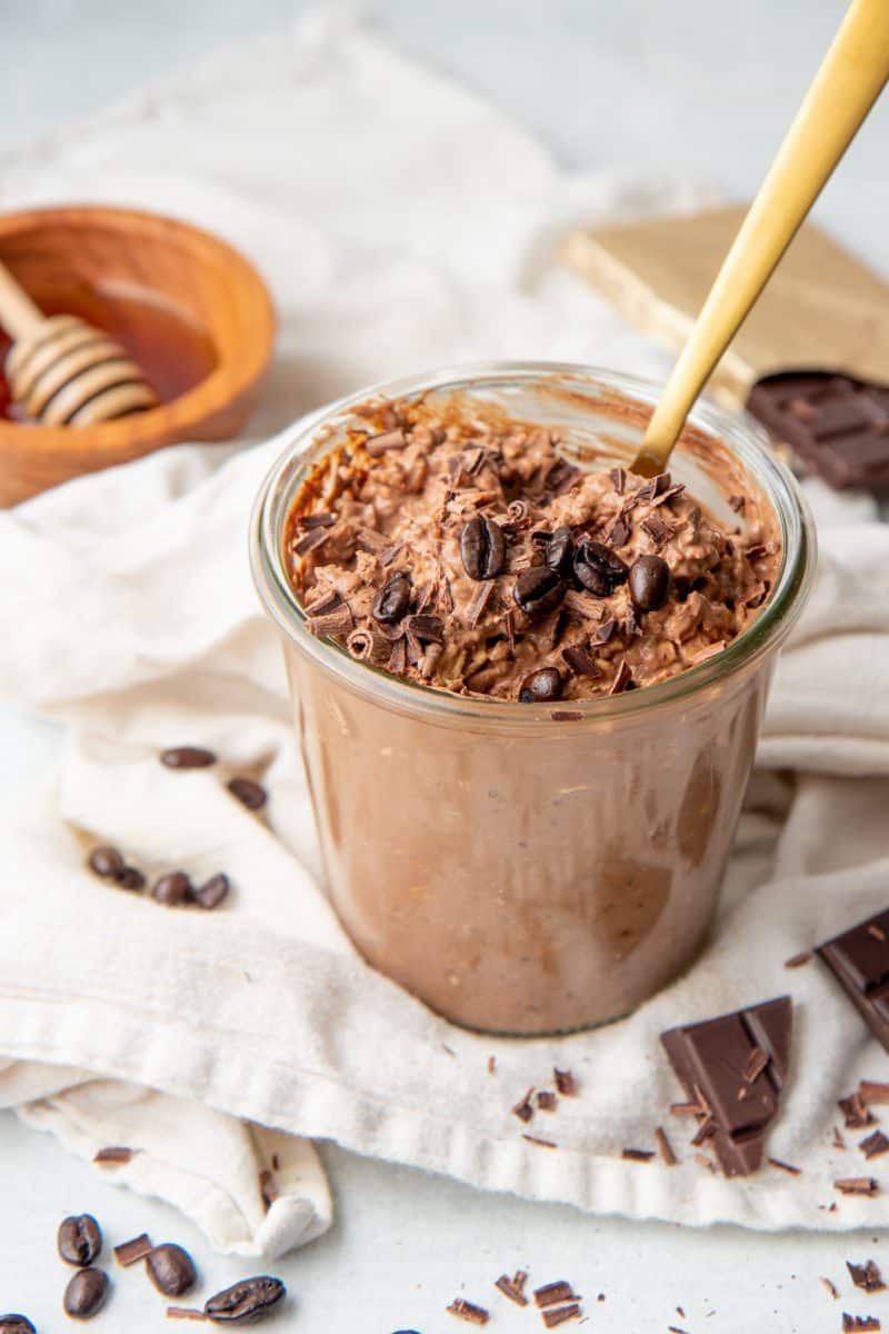 A gold spoon sits in a jar full of mocha overnight oats. The jar rests on a white dishtowel.