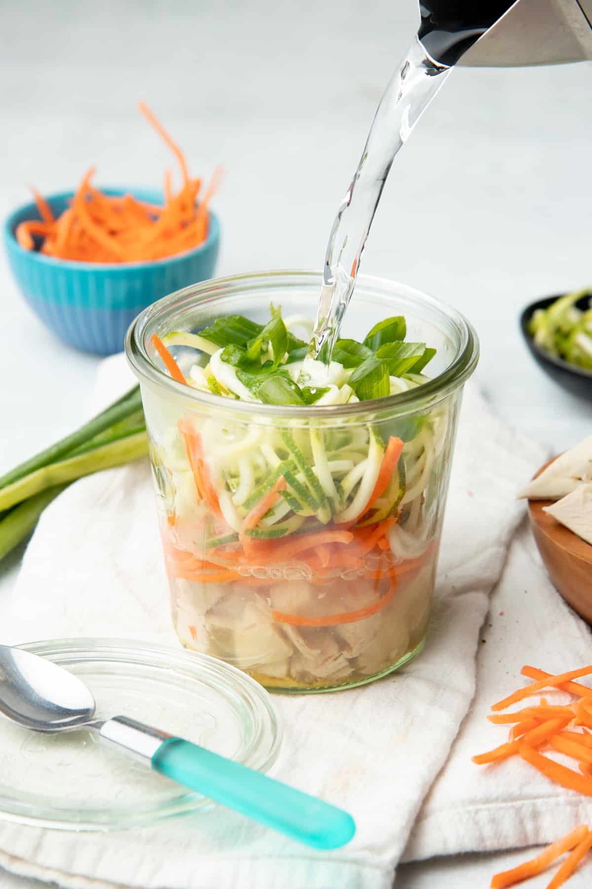 A kettle pours water into a small jar filled with chicken, vegetables, and herbs. The jar sits on a white dishtowel.