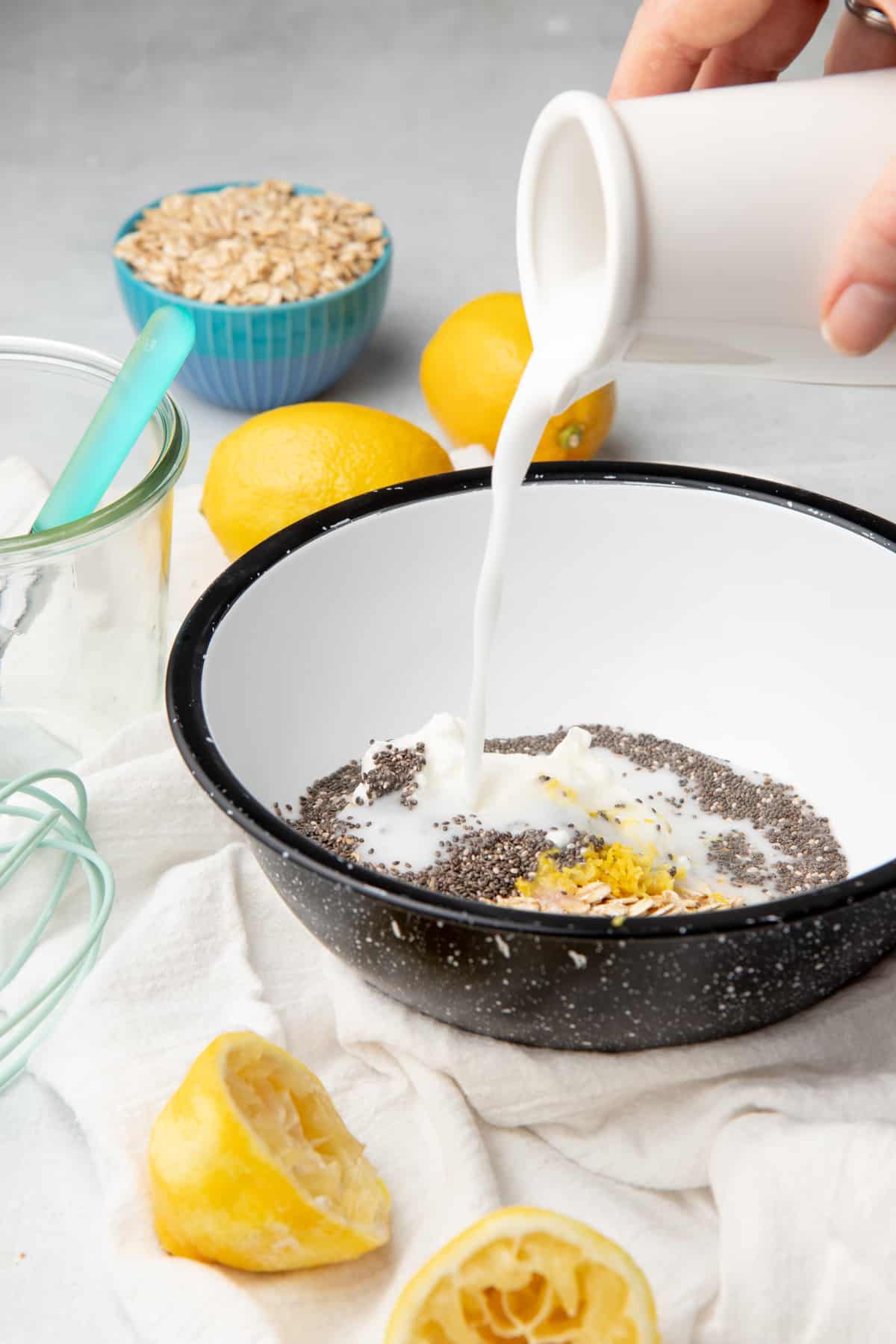 A hand pours milk onto the ingredients of overnight oats. A lemon sits in front of the bowl.