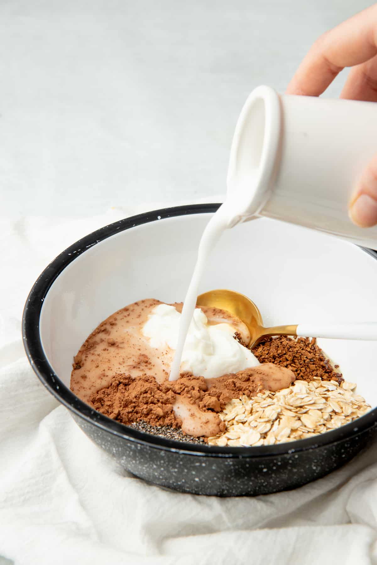 A hand pours milk into a bowl. Bowl holds oats, cocoa powder, Greek yogurt, and other ingredients.