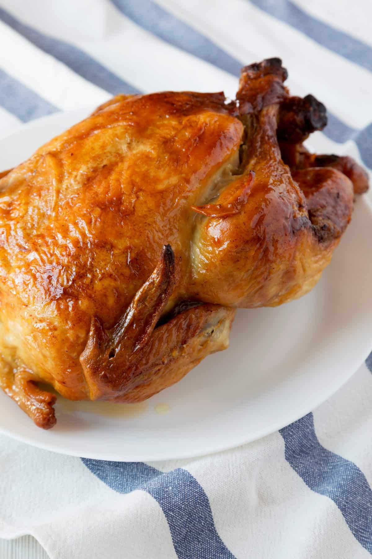 A rotisserie chicken sits on a white plate. The plate sits on a blue and white striped cloth.