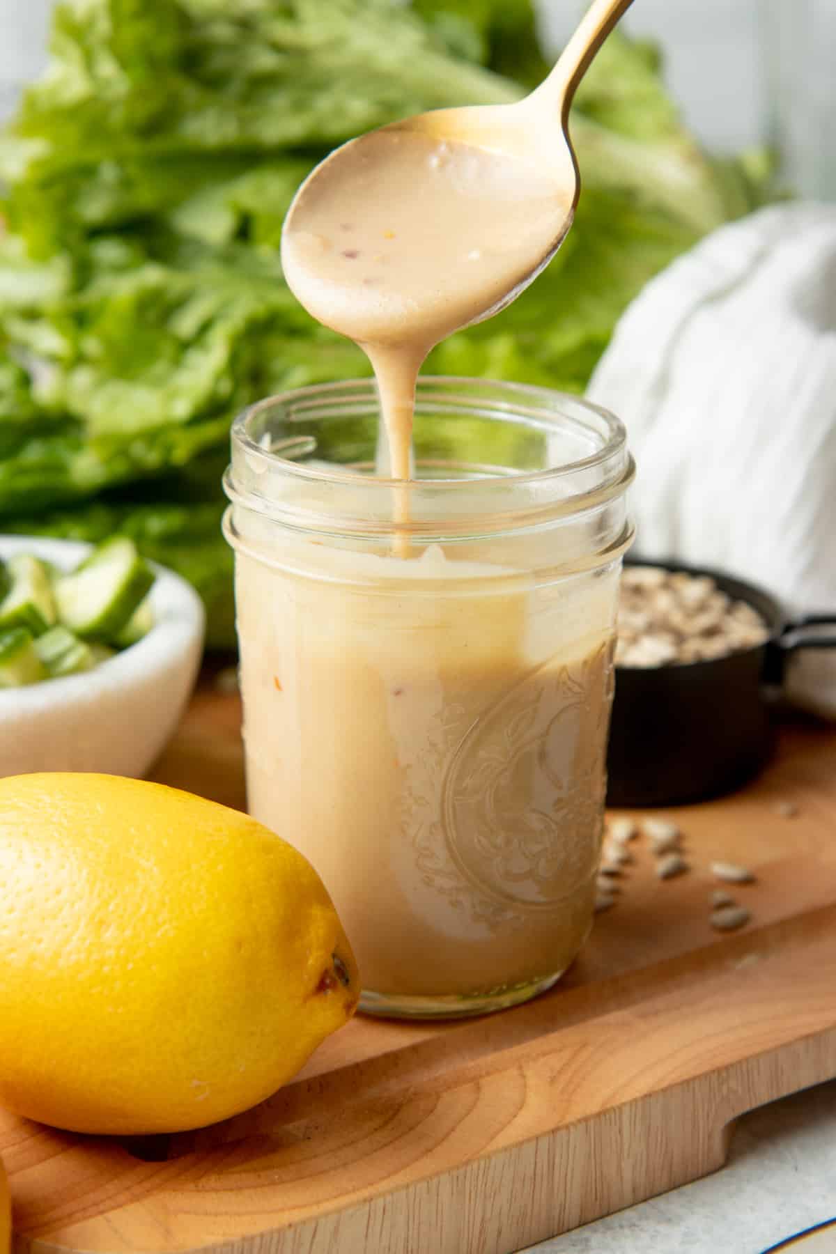 A gold spoon pours salad dressing into a mason jar sitting on a cutting board. A lemon, seeds, lettuce, and a white dishtowel are on the cutting board.