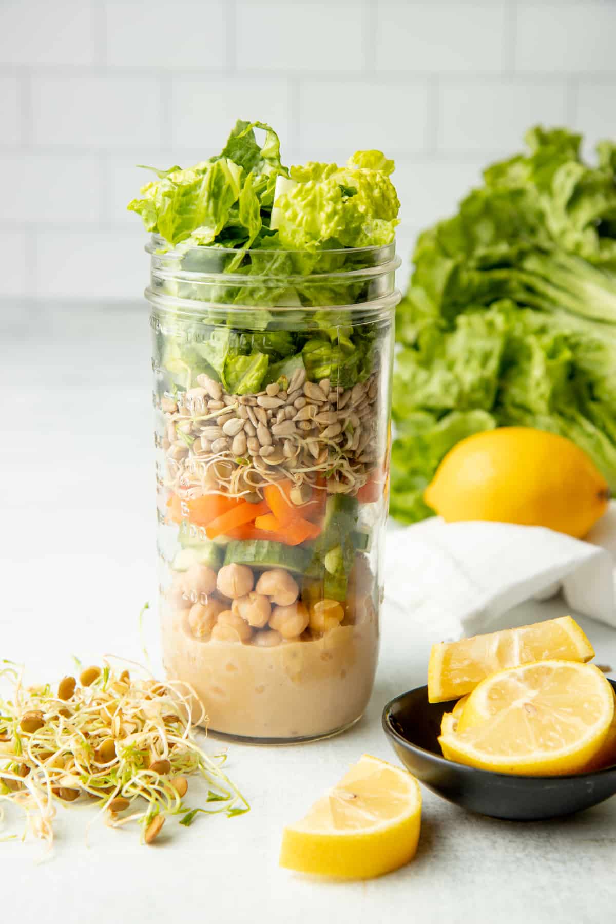 Glass mason jar filled with layers of components for a sesame chickpea salad, surrounded by extra ingredients.