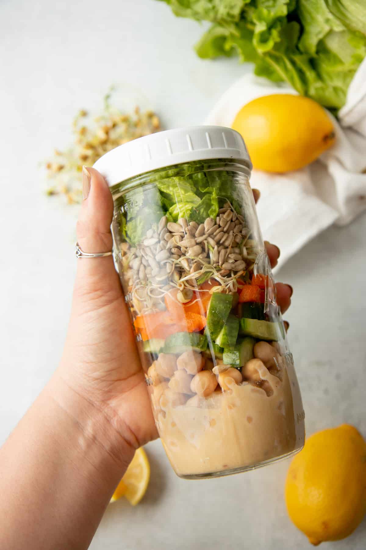 A hand holds a tall mason jar layered with salad ingredients. Lemons and lettuce rest on a white dishtowel.