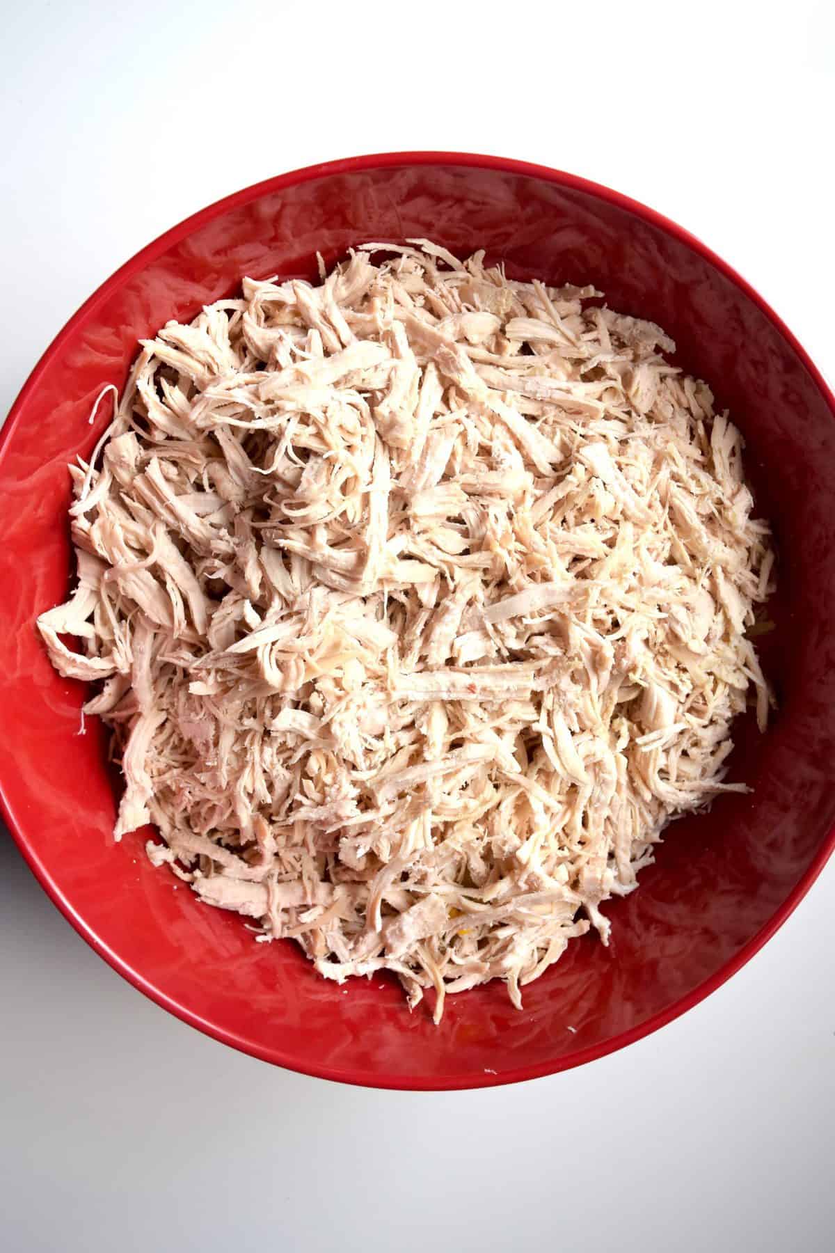 Shredded rotisserie chicken is in a large red bowl. The bowl sits on a white surface.