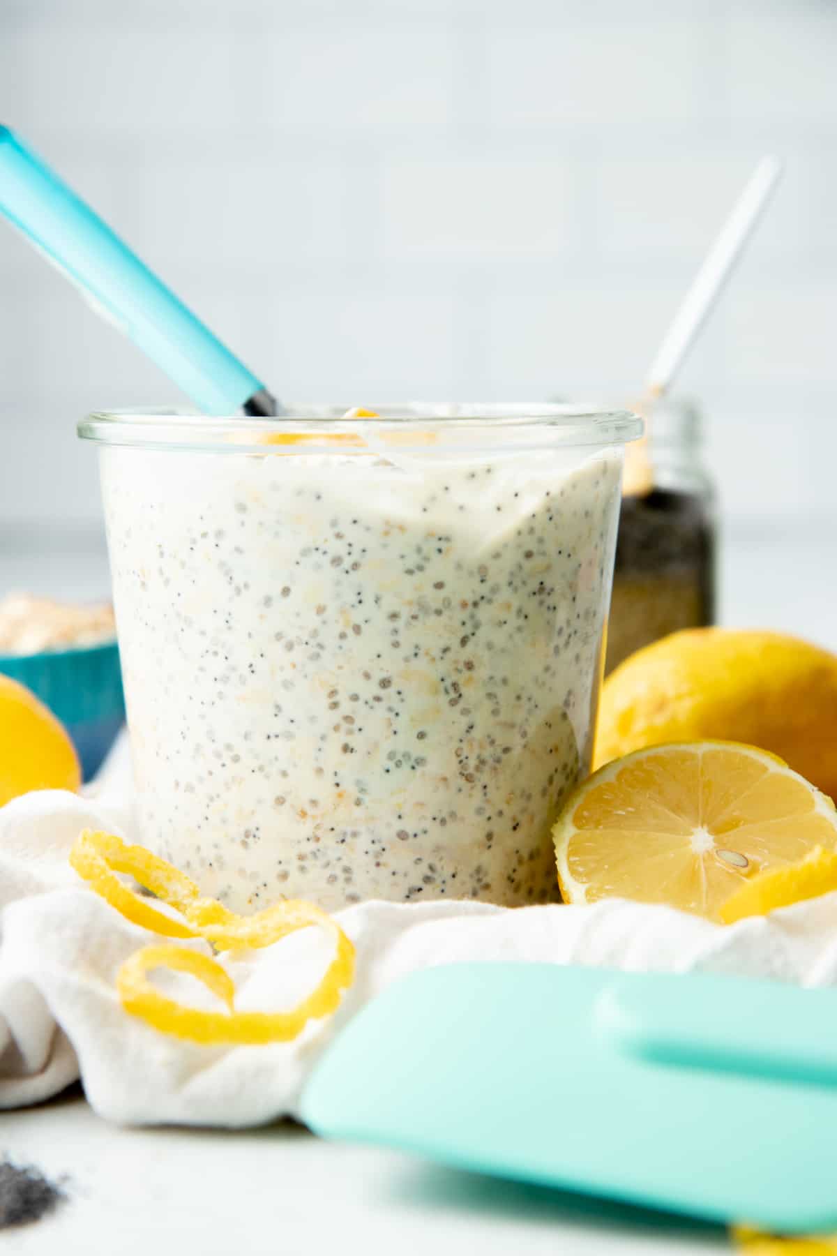 A glass jar is filled with lemon poppyseed overnight oats. Lemon and lemon zest also sit on a white dishtowel.