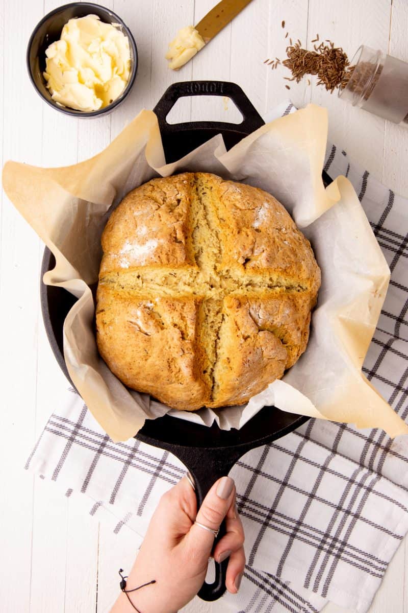 A round loaf of Irish soda bread rests in a cast iron skillet. A hand holds the handle of the pan.