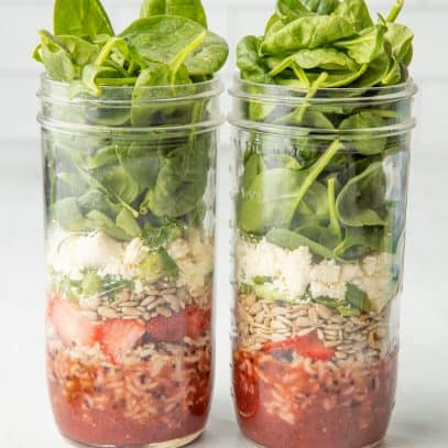 Two mason jars sit side-by-side, layered with components of strawberry spinach salads.