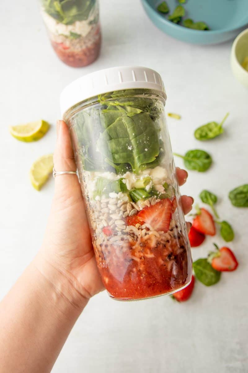 A hand holds a closed mason jar layered with the components of a strawberry spinach salad.