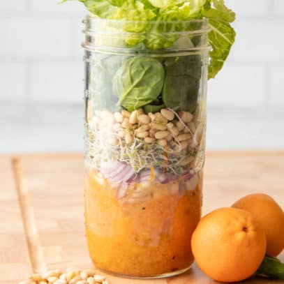 Components for a mandarin salad layered in a mason jar - dressing, mandarin slices, onions, sprouts, pine nuts, and greens. Additional ingredients surround the jar.