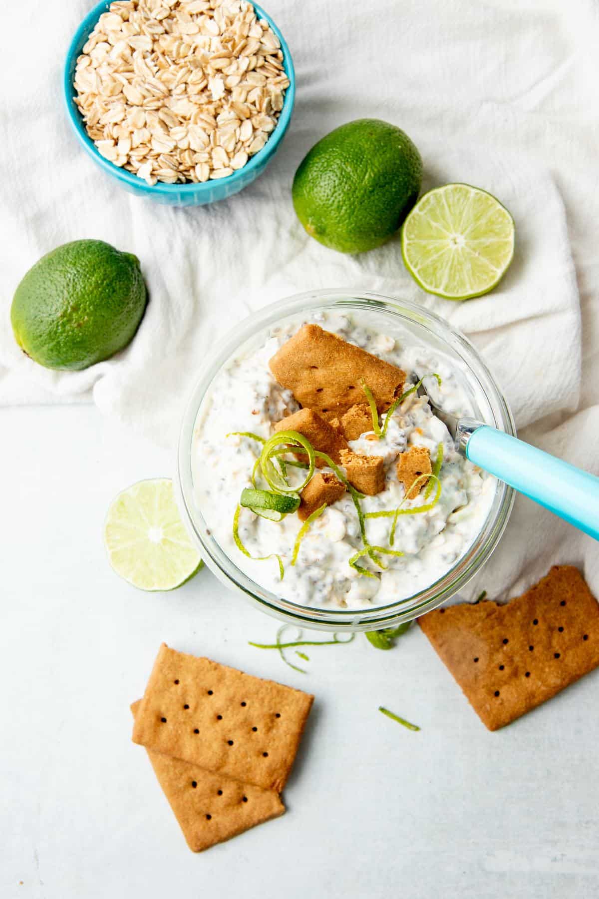 A spoon with a blue handle sits in a jar of overnight oats. Limes and graham crackers surround the jar.