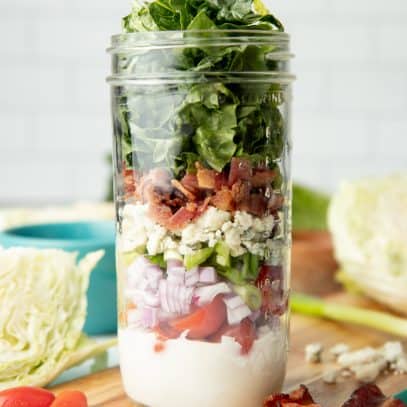 Components of a wedge salad are layered in a glass mason jar, which sits on a teal-edged cutting board. Additional ingredients surround the jar.
