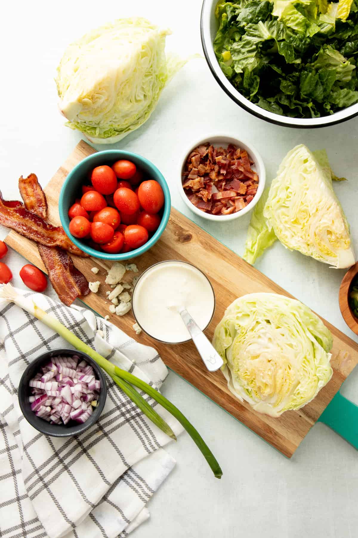 Ingredients for a wedge salad are laid out on a white counter, wooden cutting board, and plaid dish towel.
