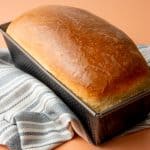 A baked loaf of bread sits in a loaf pan on top of a cloth napkin.