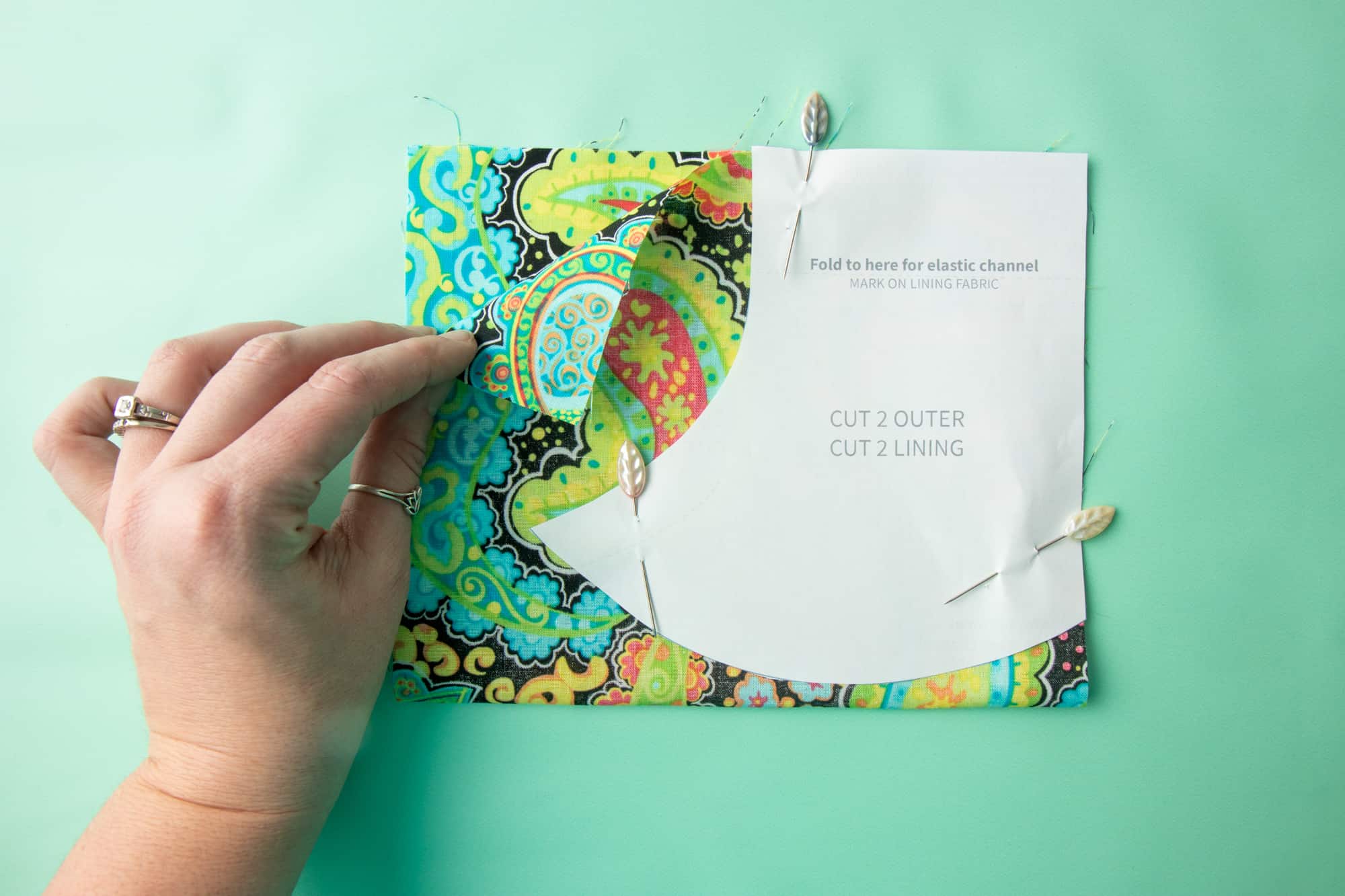 The paper pattern sits on top of the fabric, pinned, on a green background. A hand holds it in place.
