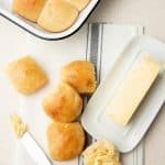Rolls lay scattered on a fabric napkin, next to a dish of butter. A knife with butter is ready to spread on one of the rolls.
