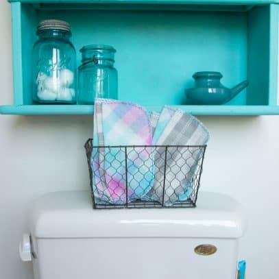 A basket of family cloth sits on top of a toilet. The wet bag hangs to the side of the toilet. A teal cabinet with mason jars on it hangs above the toilet.