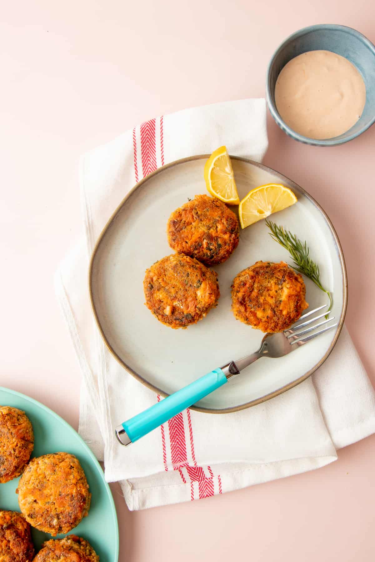 A plate of three salmon patties sits on a red and white dishcloth with a fork. A bowl of remoulade sauce sits nearby.
