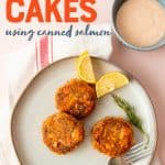 A plate of salmon cakes, lemon, and a sprig of dill sit on a plate. A bowl of Remoulade sauce sits nearby. A text overlay reads, "Salmon Cakes using canned salmon."