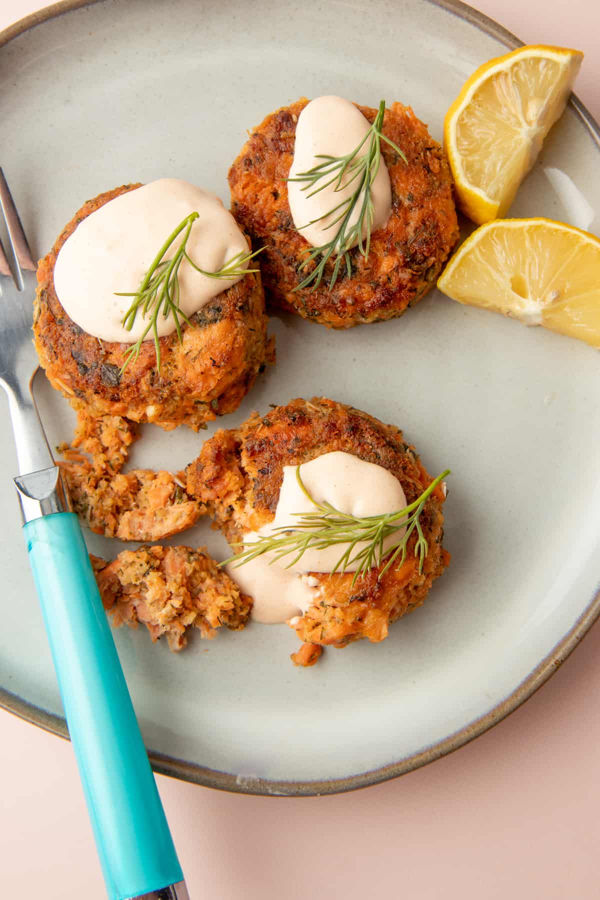 Three salmon patties dolloped with remoulade sauce and garnished with dill sit on a plate with a fork and two lemon slices.