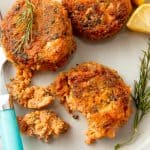 A half eaten salmon patty sits on a plate with two other cakes, a sprig of dill, and two lemon slices.