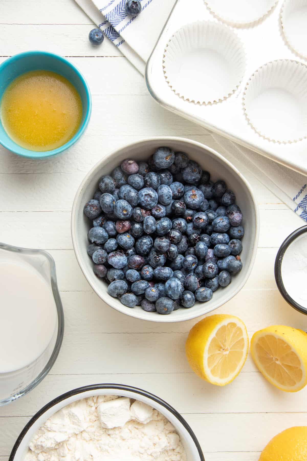 Blueberries sit in a bowl. A muffin pan, lemon slices, and other ingredients sit to the side.