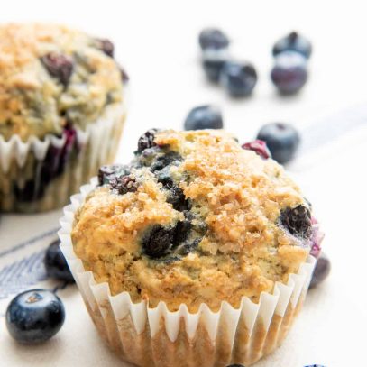 A baked Vegan Blueberry Muffin sits surrounded by fresh blueberries.