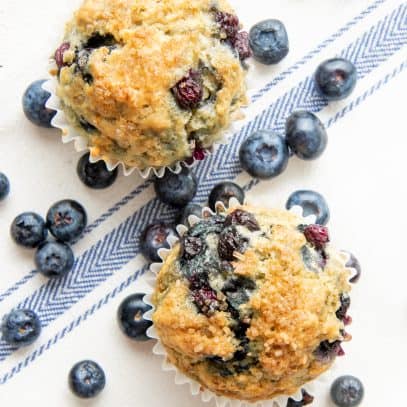 Two Vegan Blueberry Muffins sit together surrounded by fresh blueberries.